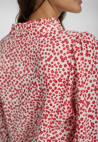 Camicia bianca con motivi floreali rossi, texture plissettata e scollatura arricciata. Il design presenta una vestibilità ampia e un tessuto morbido.