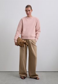 Kvinna som bär en rosa sweatshirt och vida beige byxor, håller en brun, vikt jacka, stående mot en enfärgad ljus vägg. 