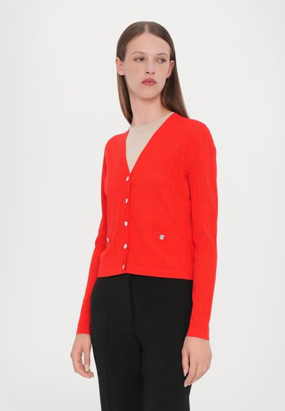 Cardigan rosso con scollatura a V, texture perforata, bottoni argentati e due tasche frontali; indossato sopra un top beige.
