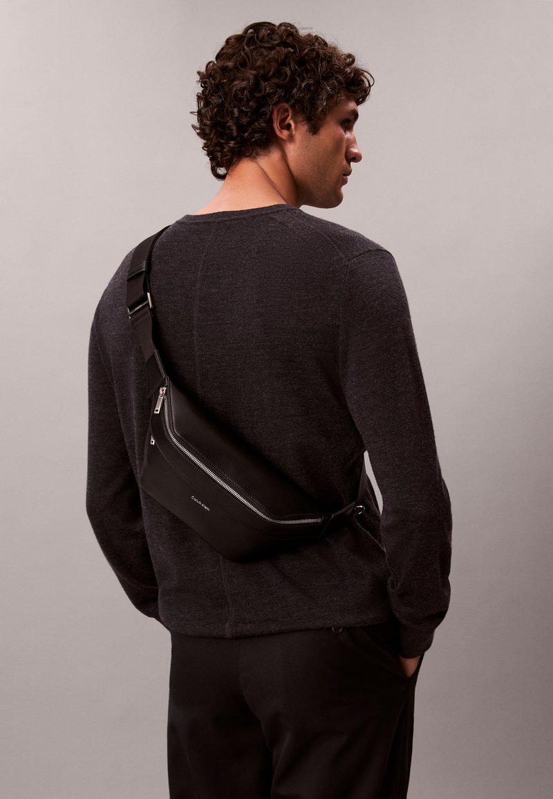 Muž s kučeravými vlasmi oblečený v tmavom svetri a nohaviciach, nesie čiernu koženú tašku cez rameno so strieborným zipsom na chrbte.