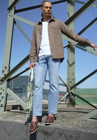 Outfit informale composto da una camicia a quadri marrone, una t-shirt bianca, jeans azzurri chiari e scarpe marroni. Gli accessori includono una borsa verde.