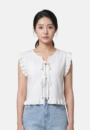 Jeune femme aux cheveux foncés attachés à l’arrière portant un haut blanc sans manches avec des volants et des liens, et un jean taille haute bleu clair.