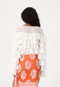Hvit gjennomsiktig topp med lagdelte volanger på ermene, kombinert med en oransje skirt med hvite paisley-mønstre og dekorative kanter.