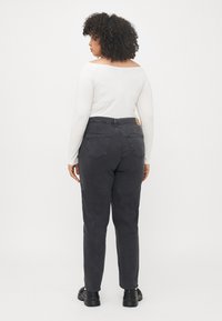 Nainen, jolla on kihara hiukset, pukeutuneena valkoiseen pitkähihaiseen puseroon, mustiin korkeavyötäisiin farkkuihin ja mustiin kenkiin, seisoo yksinkertaista valkoista taustaa vasten.