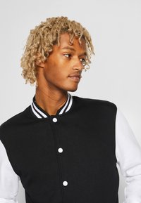 Chaqueta varsity en blanco y negro con un cuello a rayas. Presenta botones blancos y una tela texturizada. El modelo muestra un perfil lateral.