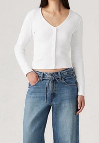 Bílý žebrovaný top s dlouhým rukávem, výstřihem do V a zapínáním na knoflíky, ve spojení s džínami s vysokým pasem, které mají na přední straně zavazovací detail.