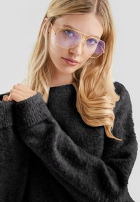 Młoda kobieta z długimi blond włosami, nosząca oversize'owe złote okulary i czarny puszysty sweter, delikatnie patrzy w kamerę.