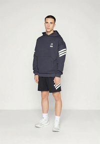 Tânăr îmbrăcat într-un hanorac bleumarin cu dungi albe și pantaloni scurți negri cu dungi albe, purtând șosete albe și adidași, pe un fundal simplu.