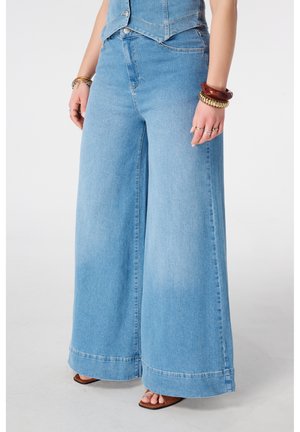 Vrouw draagt lichtblauwe jeans met wijde pijpen, bruine sandalen met open tenen en meerdere armbanden aan beide polsen, staand tegen een effen achtergrond.