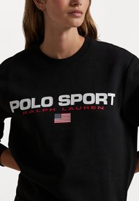 Sort sweatshirt med "POLO SPORT" og "RALPH LAUREN" i fed hvid og rød tekst, sammen med et lille amerikansk flag emblem. Blødt materiale.