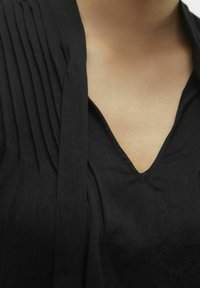 Haut noir à col en V avec des détails plissés sur les épaules et un tissu doux et texturé, présentant une coupe ample et un tombé naturel.