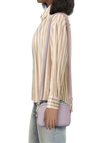 Borsa a tracolla lilla con forma strutturata, texture liscia e dettagli in ottone dorato, abbinata a una camicia beige a righe e denim chiaro.
