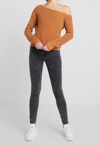Gerippter, schulterfreier, rostfarbener Pullover, kombiniert mit schwarzen Skinny-Jeans und weißen Sneakers, zeichnet sich durch eine lässige Passform und einen Texturkontrast aus.