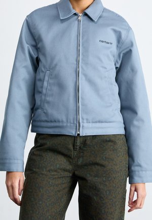 Personne portant une veste à fermeture éclair bleu clair associée à un pantalon vert foncé à motif léopard sur un fond uni.