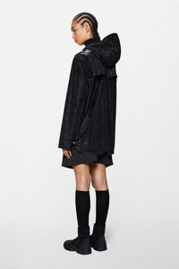 Μαύρο υπερμεγέθες μπουφάν με υφή υφάσματος, κουκούλα και περίπλοκο σχέδιο· συνδυασμένο με κοντή φούστα και μαύρες κάλτσες μέχρι το γόνατο και μπότες.