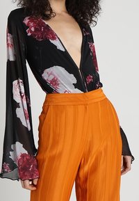 Body nero a fiori con scollatura a V profonda, maniche lunghe in tessuto trasparente; abbinato a pantaloni a vita alta in satin arancione con pieghe.