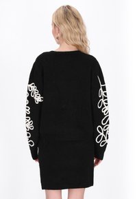 Svart tröja med långa ärmar som har broderade vita blommönster på ärmarna. Mjuk textur och avslappnad passform är tydliga.