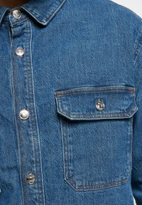 Blue denimskjorte med knapper foran, spiss krage og en brystlomme. Har metalliske trykknapper og synlige sømmer.