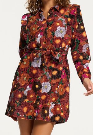 Femme portant une robe boutonnée à manches longues avec un motif floral vif orange, rose, noir et blanc, ceinturée à la taille avec une ceinture assortie.