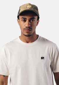 Hellbeige Baumwoll-T-Shirt mit einem kleinen schwarzen Elefantenlogo auf der Brust; kombiniert mit einer abgenutzten olivgrünen Cordmütze.