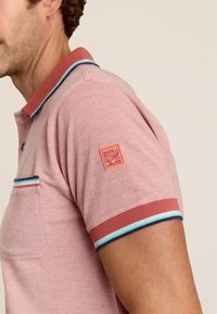 Polo en coton rose clair avec une finition texturée. Il présente un col contrasté, des poignets de manches rayés et un patch logo sur le bras.