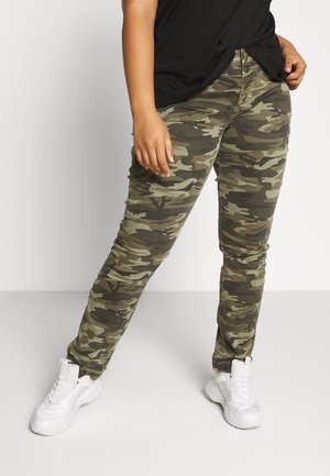 Personne portant un pantalon camouflage vert et marron, un haut noir et des baskets blanches, debout avec une jambe tendue vers l'avant.
