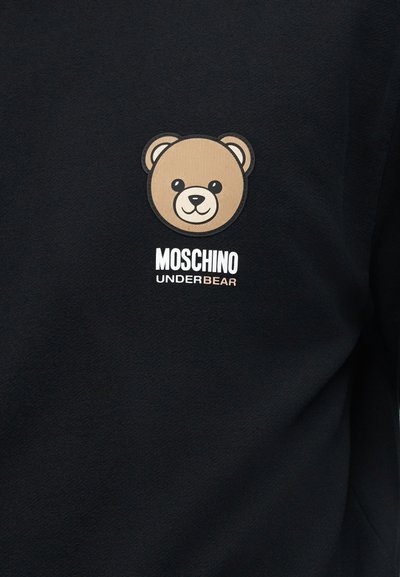 Πρόσωπο αρκουδάκι σε μπεζ χρώμα πάνω από το κείμενο «MOSCHINO UNDERBEAR» σε μαύρο ύφασμα, πιθανότατα ένα φούτερ ή μπλούζα.
