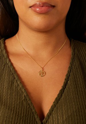 Frau mit einer goldenen Lebensbaum-Anhänger-Halskette über einem olivgrünen, strukturierten V-Ausschnitt-Top.