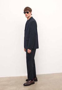 Traje de marinero a medida con blazer de un solo botón, pantalones de corte ajustado y zapatos de cuero marrón. El modelo lleva gafas de sol oscuras frente a un fondo neutro.