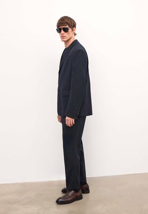 Traje de marinero a medida con blazer de un solo botón, pantalones de corte ajustado y zapatos de cuero marrón. El modelo lleva gafas de sol oscuras frente a un fondo neutro.