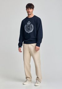 Felpa della marina con logo bianco e dettaglio della corona, abbinata a pantaloni beige chiaro e sneakers chiare. Tessuto morbido, vestibilità comoda.