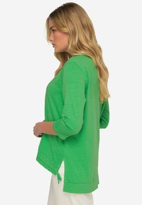 Grünes Oberteil mit 3/4-Ärmeln, das einen lockeren Schnitt und seitliche Schlitze aufweist. Der Stoff hat eine weiche Textur und ein dezentes meliertes Muster.