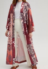 Long kimono-style outerwear in red and pink with paisley and floral patterns. Lightweight fabric, open front, wide sleeves, worn over white pants.