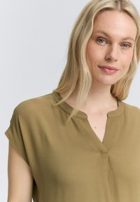 Blouse vert olive à manches courtes avec un décolleté en V et un léger froncé aux épaules. Tissu doux et texturé avec une coupe décontractée.