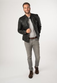 Schwarze Lederjacke mit gesteppter Schulterpartie, getragen über einem hellgrauen Hemd. Graue Jeans und braune Stiefel. Neutraler Hintergrund.