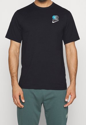 Man draagt een zwart Nike T-shirt met blauw-witte Nike Worldwide globe-logo op de linkerborst, gecombineerd met een groene broek, staand tegen een witte achtergrond.