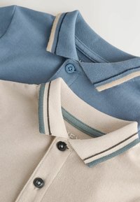 Dve zložené polokošele, jedna béžová s čiernymi gombíkmi a pruhovaným golierom, a jedna modrá s farebne zladenými gombíkmi a pruhovaným golierom.