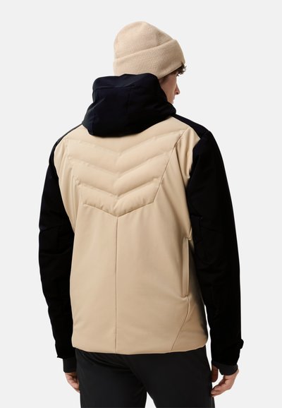 Beige en zwarte jas met een gewatteerd chevronpatroon op de rug, met een capuchon en zwarte mouwen. Gehaakte beige beanie op het hoofd.