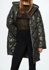 Donna che indossa un cappotto imbottito a sacco verde scuro sopra un top trasparente a strisce nere e marroni, con pantaloni neri, in piedi con una mano nella tasca.