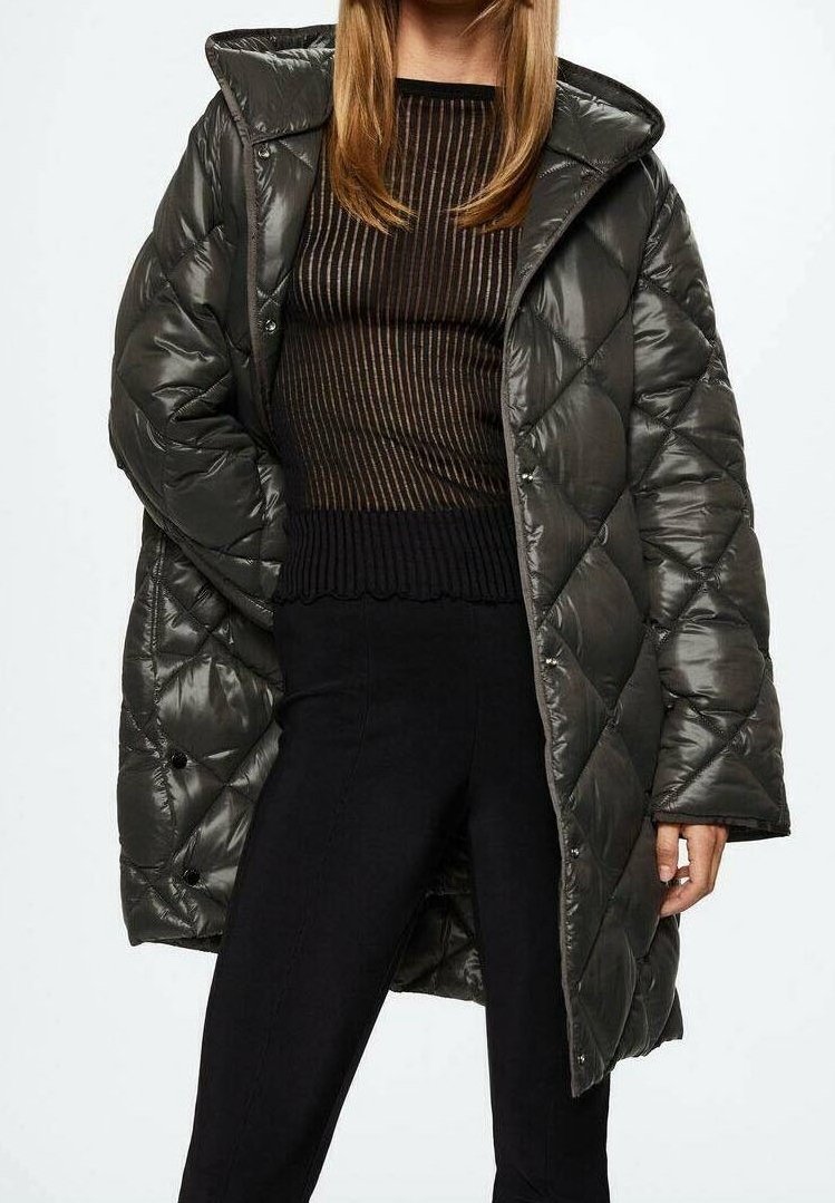 Donna che indossa un cappotto imbottito a sacco verde scuro sopra un top trasparente a strisce nere e marroni, con pantaloni neri, in piedi con una mano nella tasca.