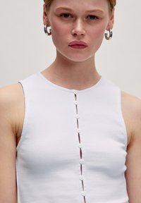 Weißes, geripptes Tanktop mit rundem Ausschnitt, das vorne mit kleinen weißen Knöpfen versehen ist. Der Stoff wirkt glatt und enganliegend.