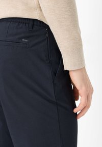 Pantaloni blu navy con tasca posteriore con zip, realizzati in tessuto liscio. Un maglione beige lavorato a maglia copre parte della mano appoggiata sull'anca.