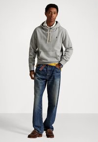 Grå huvtröja med dragsko, stor framficka och broderad logotyp, kombinerad med urblekta blå jeans och bruna skor.