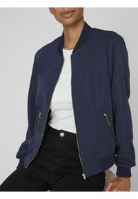 Mörkblå bomberjacka med guldfärgade blixtlåsdetaljer, ribbade muddar och midjeband, lätt tyg, kombinerad med en vit skjorta och svarta byxor.