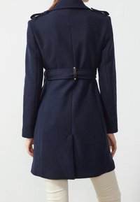 Kobieta w ciemnonawigacyjnej, zapinanej na pasek płaszczu z epauletami, pokazana z tyłu, w zestawieniu z jasnobeżowymi spodniami na jednolitym tle.