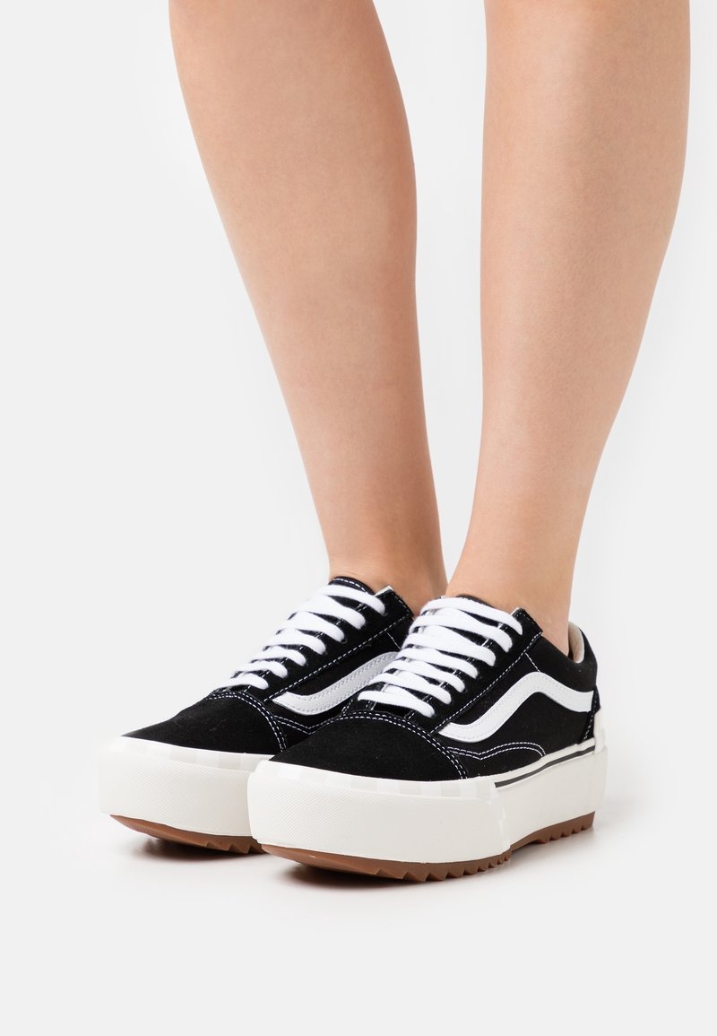 Pieds portant des baskets basses noires et blanches avec des semelles blanches épaisses et des lacets blancs sur fond blanc.