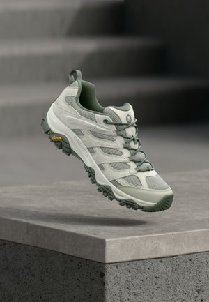 Világosszürke és zöld túracipő Vibram talppal, amely egy beton tömb felett lebeg, elmosódott lépcsős háttér előtt.