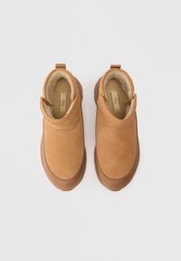 Barna velúr slip-on cipők puha, bolyhos béléssel, lekerekített orral, gumitalppal és egy húzófüllel a sarkán a könnyű felhúzás érdekében.