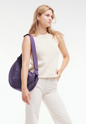 Lila Nylon Schultertasche mit großer, runder Form, verstellbarem Riemen und minimalistischem Design, getragen über einem hellen Oberteil und einer Hose.