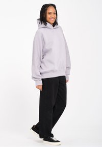 Haut de hoodie surdimensionné violet clair avec capuche à cordon, poignets et ourlet côtelés, associé à un pantalon large noir et des baskets noires.